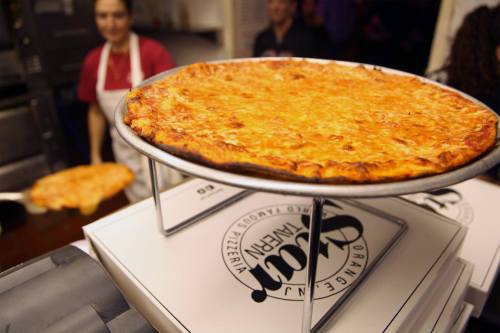 A large, cheesy pizza on a metal stand, with a pizzeria backdrop and staff in the background.
