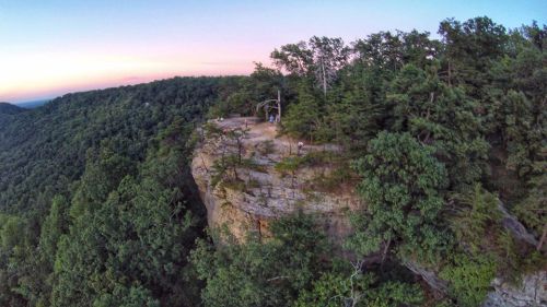Aerial view of a rocky cliff surrounded by lush trees at sunset, with people enjoying the scenic overlook.