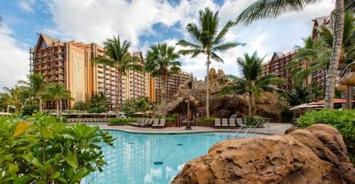 Tropical resort pool surrounded by palm trees and lush greenery, with large hotel buildings in the background.