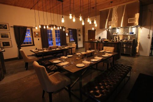 A cozy dining room with wooden tables, elegant chairs, and warm lighting, featuring a modern kitchen in the background.