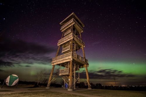A wooden observation tower under a starry sky with vibrant green auroras illuminating the night.