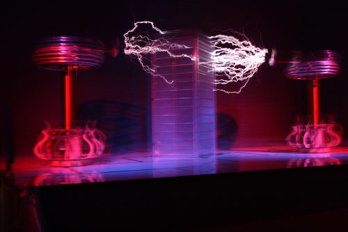 A dramatic display of electricity arcs between two Tesla coils, illuminating a dark stage with vibrant colors.