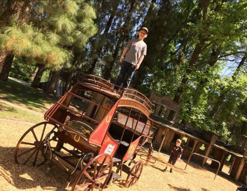 A child stands on top of a vintage wagon in a playground, surrounded by trees and another child playing nearby.