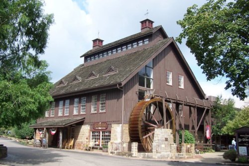 A large, rustic wooden building with a water wheel, surrounded by trees and a cloudy sky.
