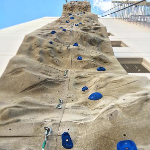 A tall rock climbing wall with blue holds against a textured surface, reaching up towards a clear blue sky.