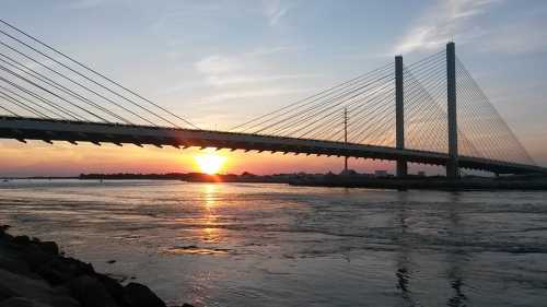 A cable-stayed bridge spans a river at sunset, with vibrant colors reflecting on the water.