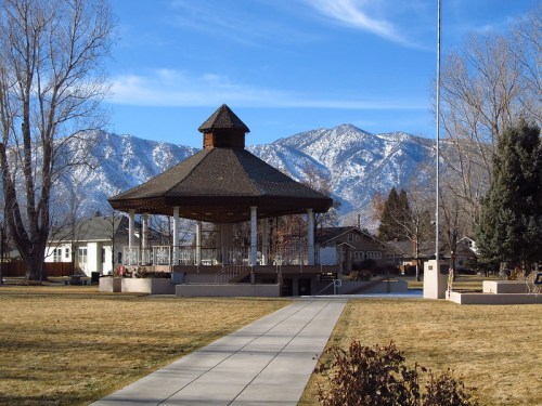 A gazebo in a park with mountains in the background, under a clear blue sky and surrounded by grass and trees.