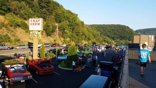 A car show with various classic cars and spectators, set against a scenic hillside and a diner sign in the background.