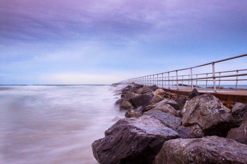 A long pier extends over rocky shoreline, with gentle waves lapping against the stones under a cloudy sky.
