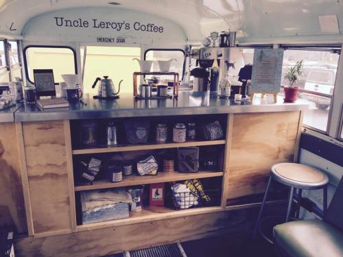Interior of a coffee bus with a counter, coffee equipment, and shelves displaying jars and supplies. Cozy atmosphere.