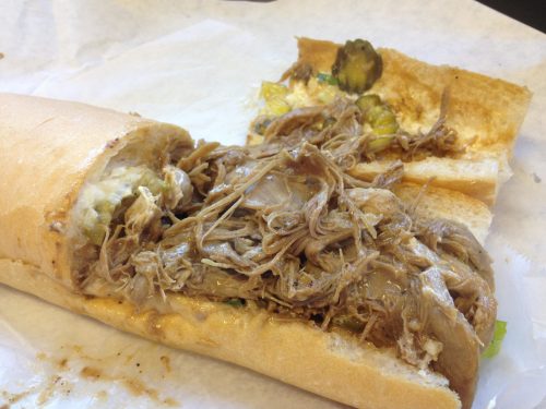 A close-up of a sandwich filled with shredded meat, pickles, and lettuce on a toasted hoagie roll.