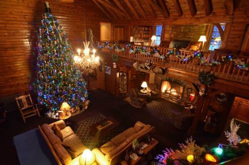Cozy log cabin interior decorated for Christmas, featuring a large tree, fireplace, and festive lights.