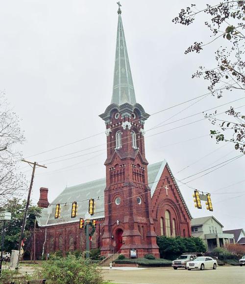 A tall, red brick church with a spire, surrounded by trees and power lines, on a cloudy day.