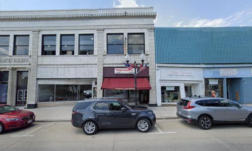 A street view featuring two storefronts, one with a red awning and a parked gray SUV in front.
