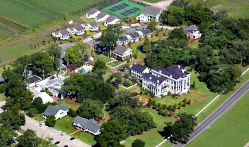 Aerial view of a residential area with multiple houses, a pool, tennis court, and surrounding greenery.