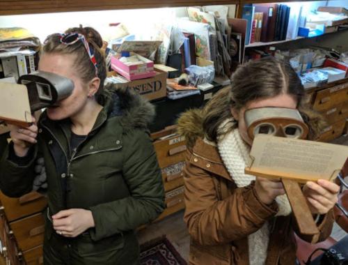 Two women using vintage viewers in a cluttered room filled with books and artifacts.
