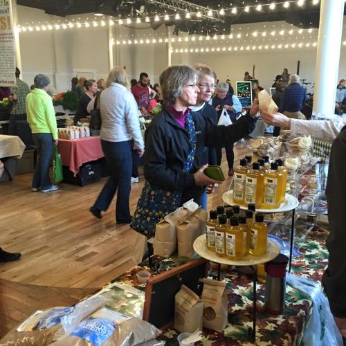 A bustling market scene with shoppers browsing stalls filled with products, including oils and baked goods.