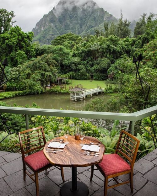 A scenic view from a balcony featuring a wooden table set for two, overlooking lush greenery and a serene pond.