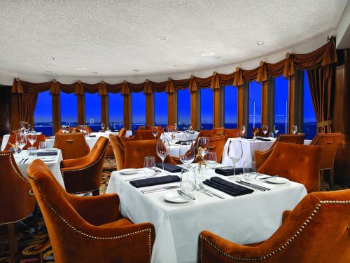 Elegant restaurant interior with round tables, plush chairs, and panoramic windows showcasing a night view.