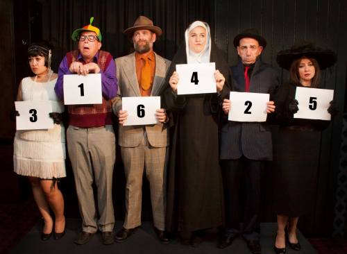 A group of six people in costumes holding numbered signs, posing in a lineup against a dark background.