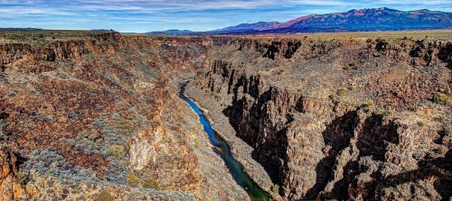 A scenic view of a deep canyon with a winding river, surrounded by rugged cliffs and distant mountains under a clear sky.