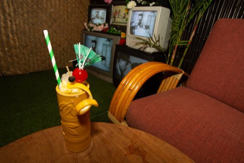 A colorful tropical drink with a cherry and lemon garnish sits on a table next to a retro chair and vintage TVs.