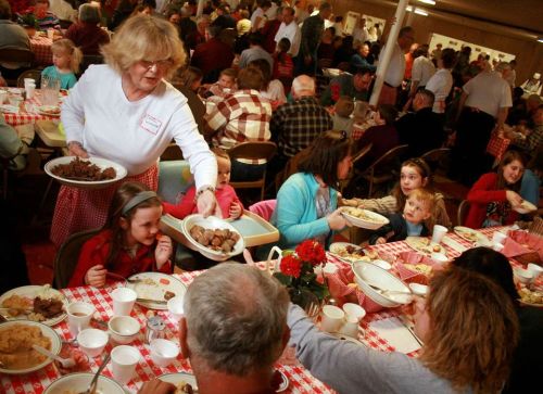 A busy community gathering with people enjoying a meal, as a woman serves food to children at a table.