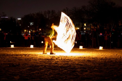 A performer spins a flaming object in a snowy park at night, surrounded by spectators and glowing lanterns.