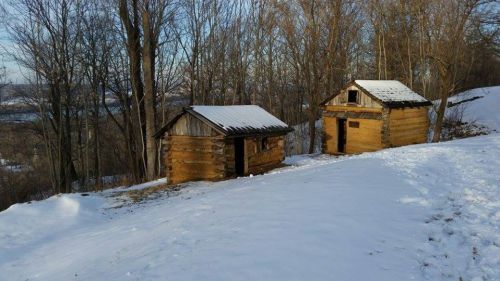 Two rustic wooden cabins sit on a snowy hillside, surrounded by bare trees and a clear blue sky.