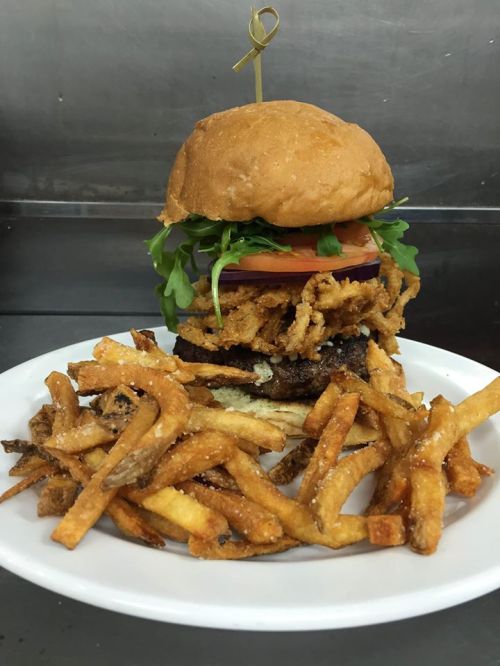 A towering burger with lettuce, tomato, and crispy onions, served with a side of golden French fries on a white plate.