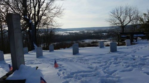 A snowy landscape with gravestones and small American flags, overlooking a valley under a clear sky.