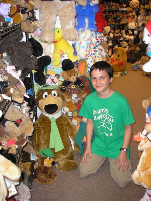 A child in a green shirt kneels beside a large stuffed dog in a store filled with various plush toys.