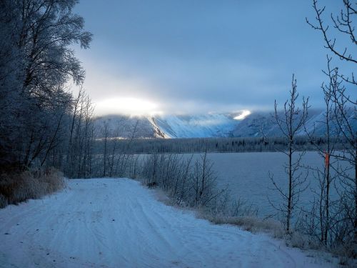 A snowy path leads to a serene lake, surrounded by frosty trees and mountains under a cloudy sky.