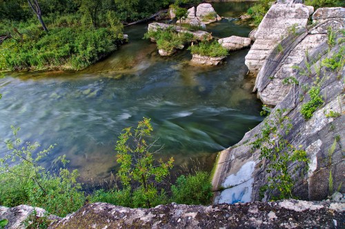 A serene river flows between rocky banks, surrounded by lush greenery and trees under a clear sky.