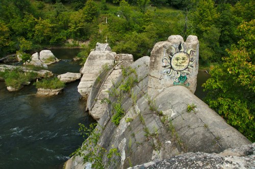 A river flows past weathered concrete remnants, featuring a sun mural, surrounded by lush greenery.