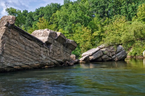Rugged rock formations emerge from calm water, surrounded by lush green trees under a partly cloudy sky.