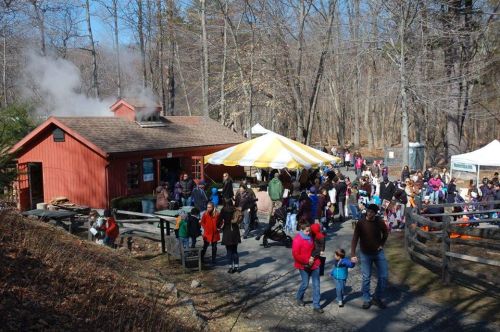 A crowd gathers outside a red building with smoke rising, surrounded by trees and a striped tent in a park setting.
