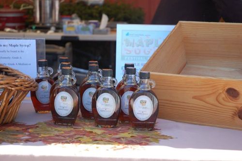 Bottles of maple syrup displayed on a table with autumn leaves and informational signs at a festival.