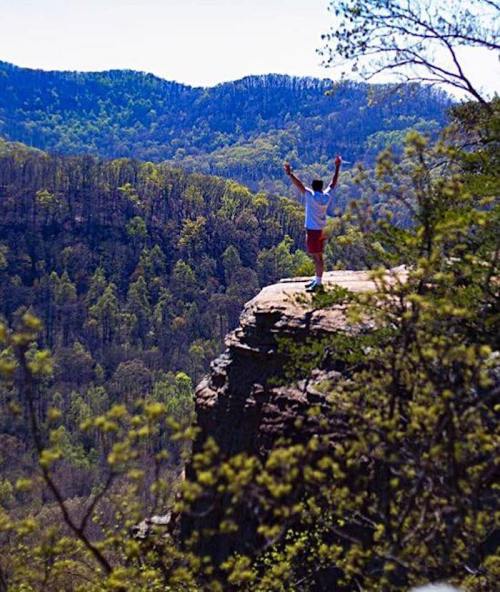 A person stands on a rocky ledge, arms raised, overlooking a lush green valley and distant mountains.