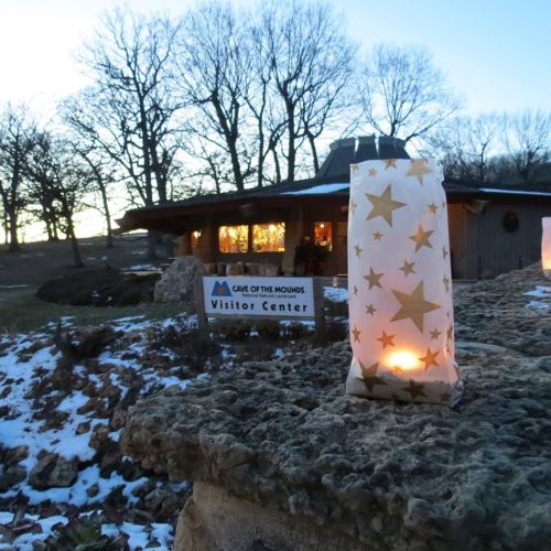 A lit lantern with stars sits on a rock in front of the Cave of the Mounds Visitor Center at dusk. Snow is visible.