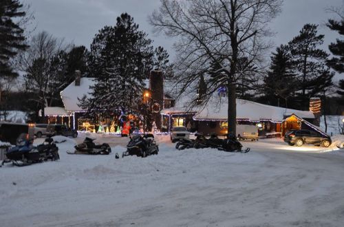 A snowy scene featuring a decorated house with holiday lights and parked snowmobiles in a winter landscape.