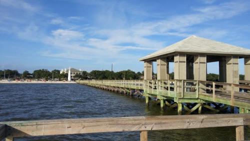 A wooden pier extends over calm water, leading to a gazebo, with a beach and trees in the background under a blue sky.