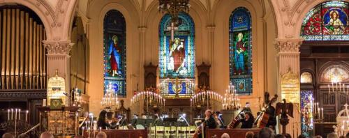 A musical performance in a church with stained glass windows and candlelight, featuring musicians and an orchestra.