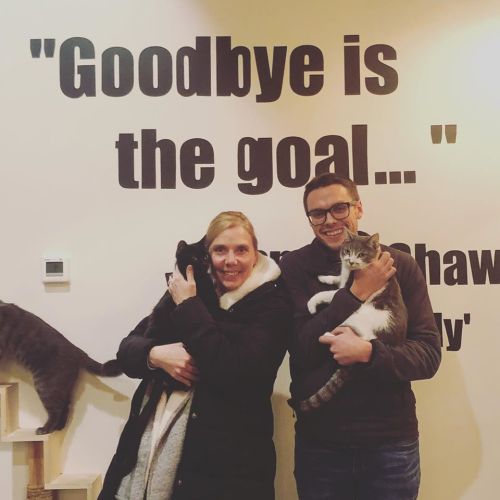 A man and a woman hold cats, standing in front of a wall with the quote "Goodbye is the goal..." in a cozy setting.