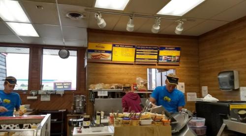 Two employees in blue shirts prepare food in a casual eatery with a wooden interior and a menu displayed above.