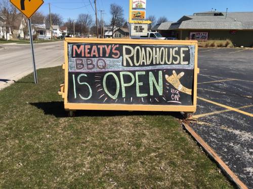 A colorful sign outside a BBQ roadhouse reads "Meaty's BBQ Roadhouse is OPEN" with a playful design.