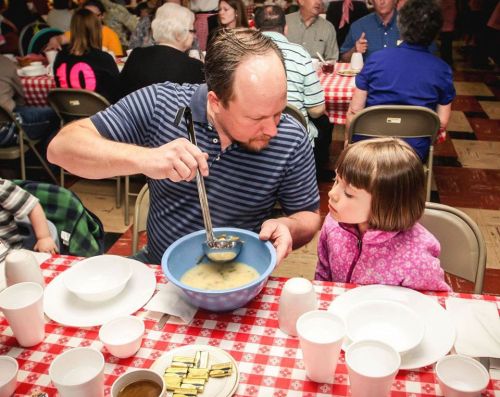A man serves soup from a bowl while a young girl watches attentively at a communal dining event.