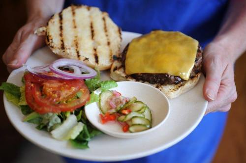 A person holds a grilled cheeseburger with a side salad and pickles on a plate.