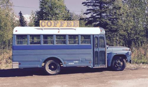 A blue vintage bus converted into a coffee stand, parked in a forested area with a "COFFEE" sign on top.