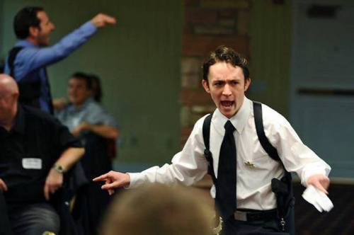 A man in a suit passionately gestures while speaking, with others engaged in conversation in the background.